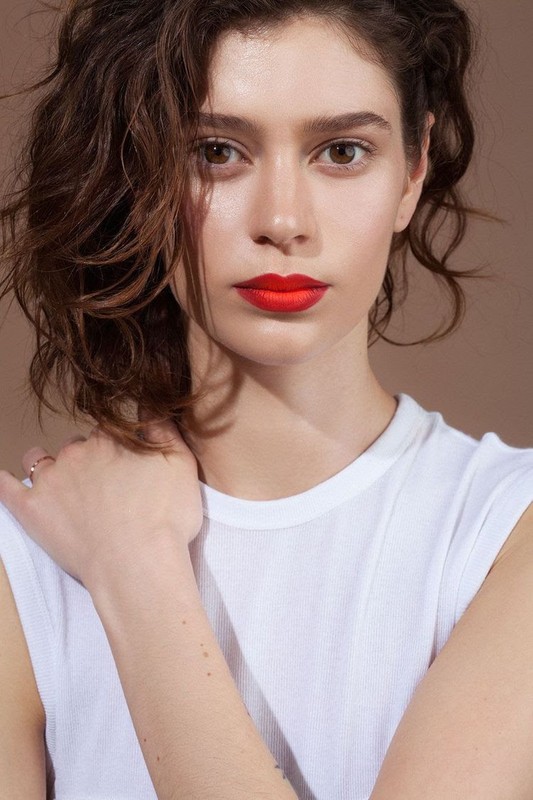 Close-up portrait of a woman with dark curly hair wearing a white top and vibrant red lipstick against a plain brown background.