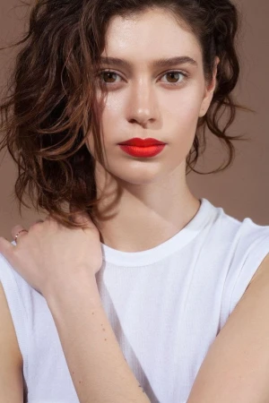 Close-up portrait of a woman with dark curly hair wearing a white top and vibrant red lipstick against a plain brown background.
