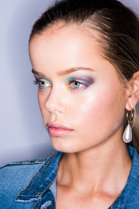 Close-up beauty portrait of a woman wearing blue and purple eyeshadow and a denim jacket with a pearl drop earring.