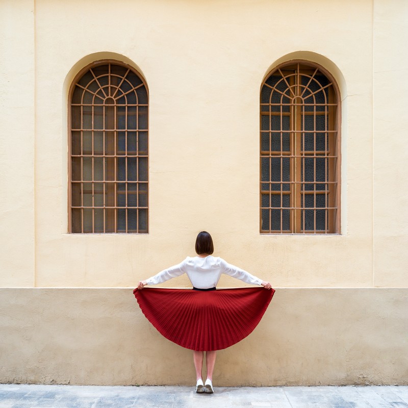 Back view of a woman holding a red pleated skirt, standing in front of a beige wall with two arched windows, fashion photography.