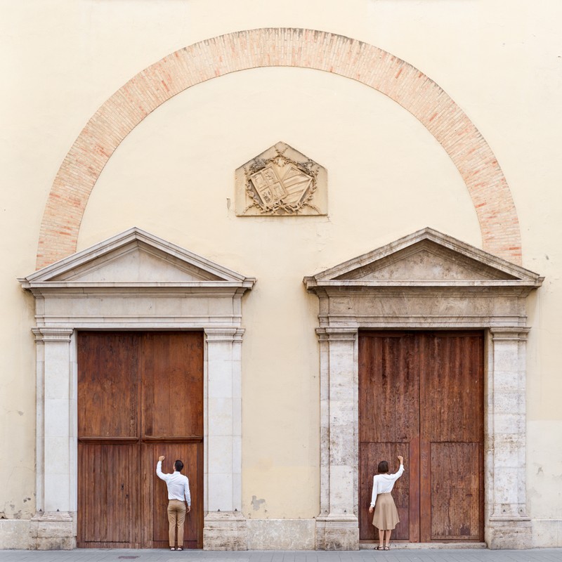 A man and a woman stand at separate wooden doors under an archway on a building, reaching up to knock, photographed at eye level.