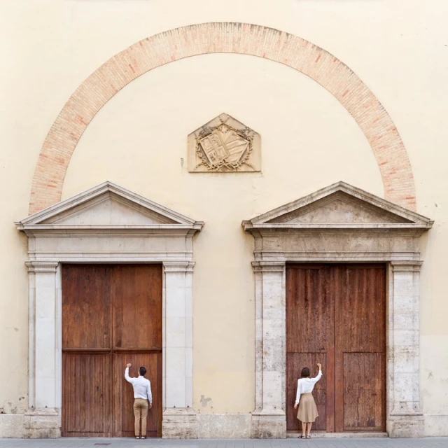 A man and a woman stand at separate wooden doors under an archway on a building, reaching up to knock, photographed at eye level.