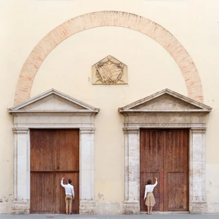 A man and a woman stand at separate wooden doors under an archway on a building, reaching up to knock, photographed at eye level.