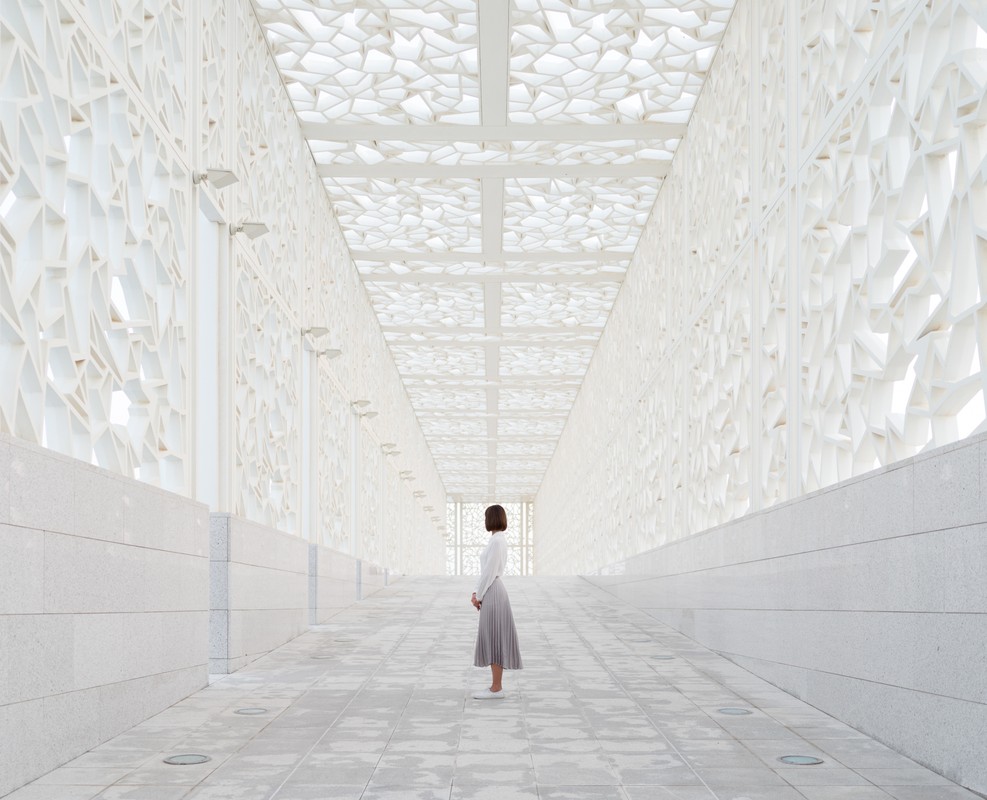 Woman in a long grey skirt stands in an all-white hallway with geometric designs on walls and ceiling, facing into the distance.