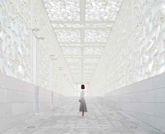 Woman in white top and grey pleated skirt standing inside a white geometric architectural walkway with patterned ceiling