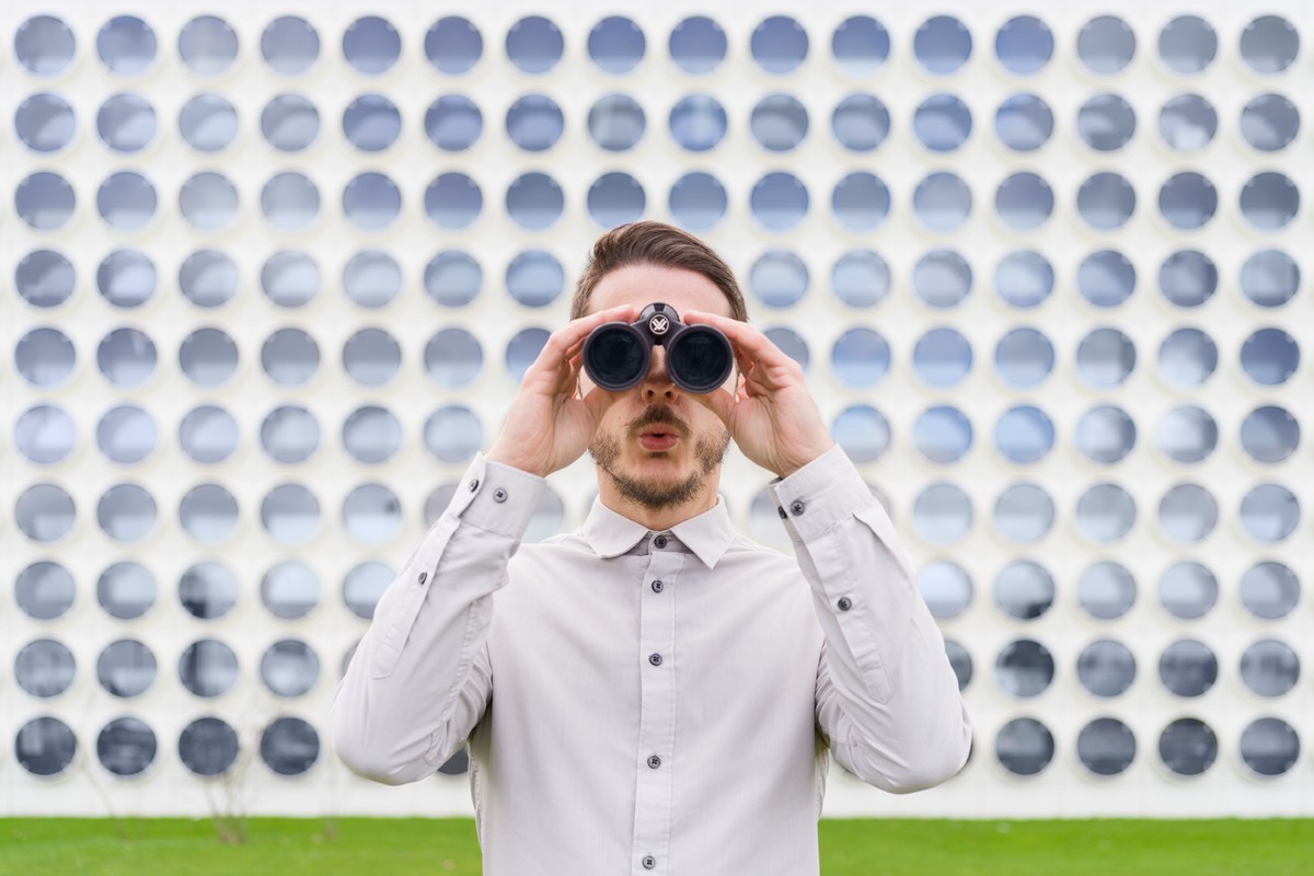Adult male looking through binoculars outdoors against a wall with repeating circular patterns in front of green vegetation.