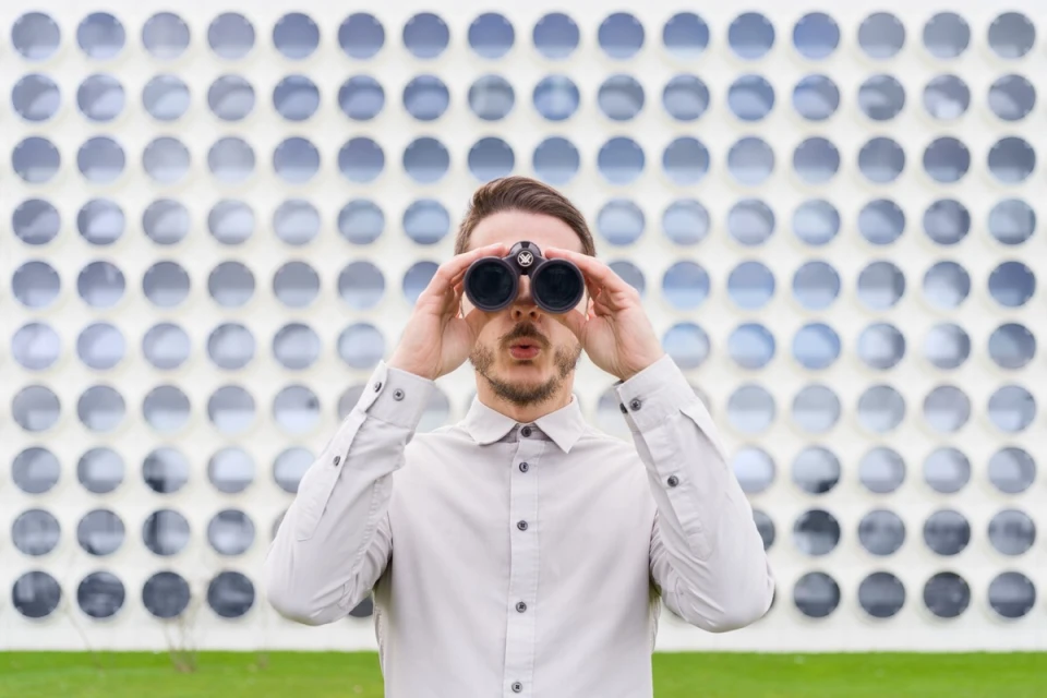 Adult male looking through binoculars outdoors against a wall with repeating circular patterns in front of green vegetation.