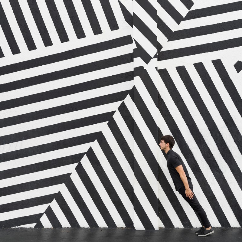 Adult male in black clothing poses against a black and white striped wall, creating a visually striking geometric pattern and illusion.
