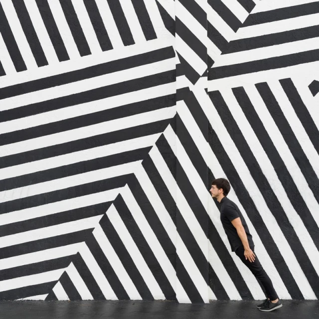 Adult male in black clothing poses against a black and white striped wall, creating a visually striking geometric pattern and illusion.