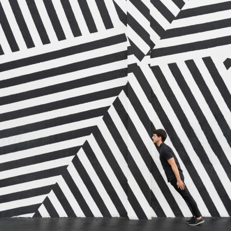Adult male in black clothing poses against a black and white striped wall, creating a visually striking geometric pattern and illusion.