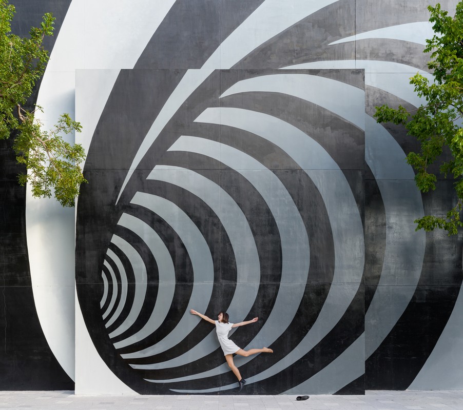 A woman in a white dress leaps in front of a black and grey abstract spiral mural outdoors on a sunny day, framed by greenery.