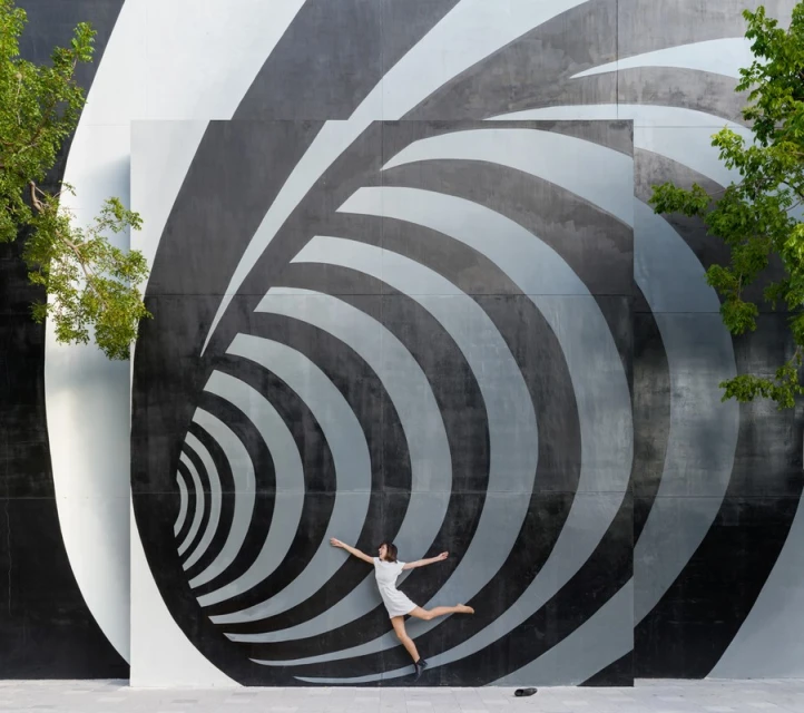 A woman in a white dress leaps in front of a black and grey abstract spiral mural outdoors on a sunny day, framed by greenery.
