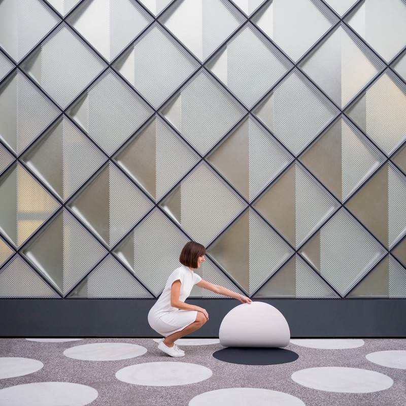Elegant architectural photo features a woman in white dress interacting with a modern, spherical seat against a patterned wall.
