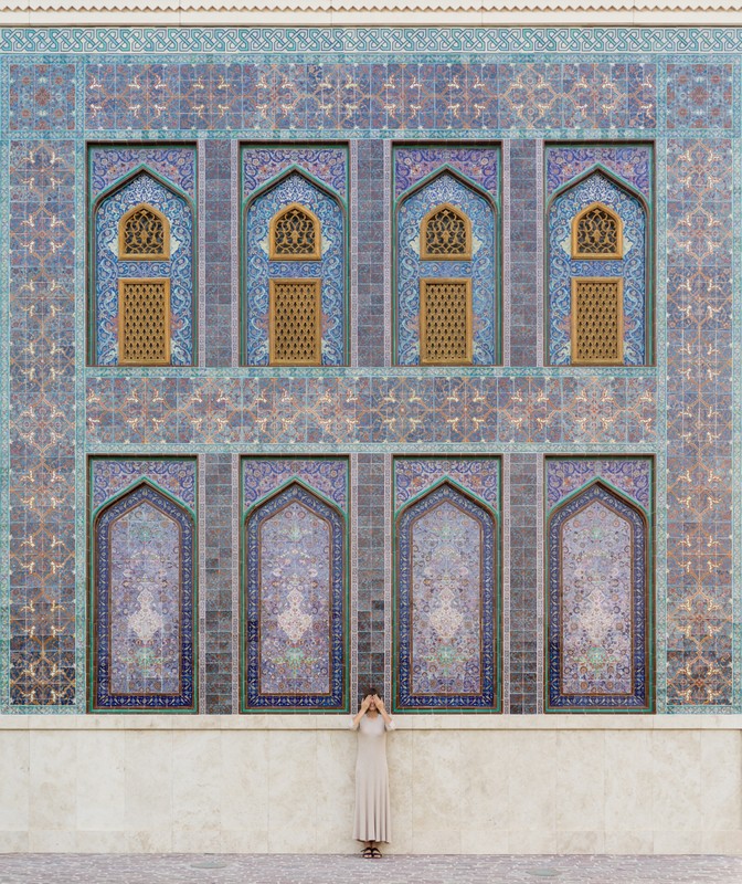 Woman in long dress stands against an intricately patterned building facade with arches, geometric designs, and muted blue and brown tones.