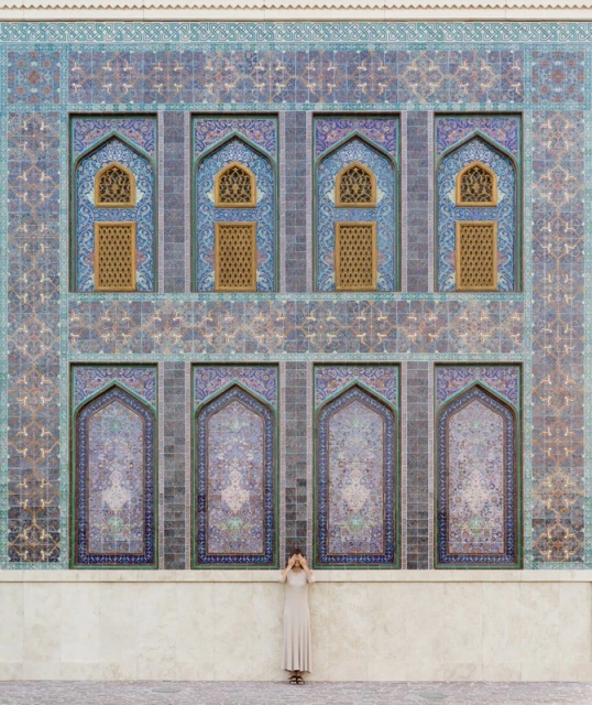 Woman in long dress stands against an intricately patterned building facade with arches, geometric designs, and muted blue and brown tones.