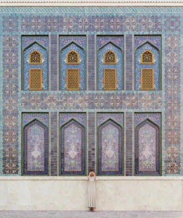 Woman in long dress stands against an intricately patterned building facade with arches, geometric designs, and muted blue and brown tones.