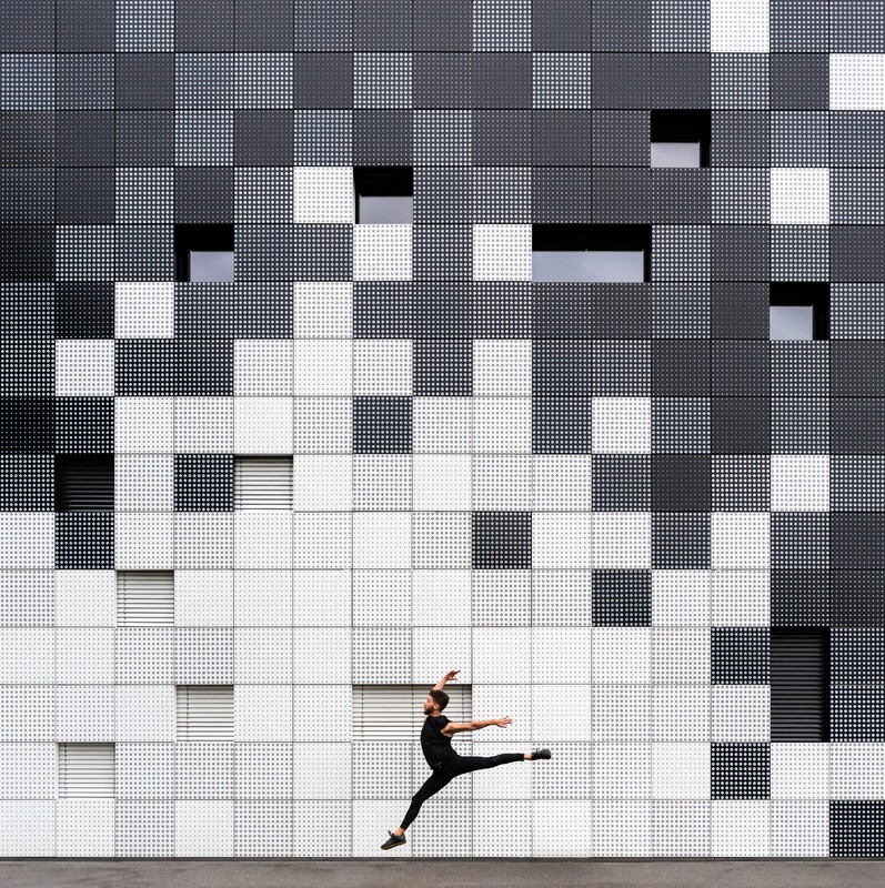 Male dancer in mid-air leap against a modern black, gray, and white building façade with geometric patterns and strategically placed windows.