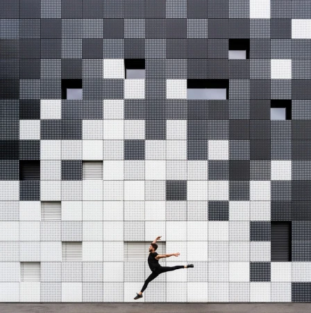 Male dancer in mid-air leap against a modern black, gray, and white building façade with geometric patterns and strategically placed windows.