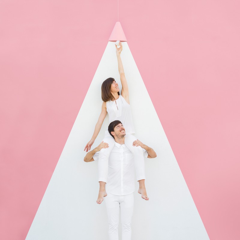Woman on man's shoulders reaches for bulb, both wearing white, against a pink and white geometric backdrop, lit by hanging lamp.