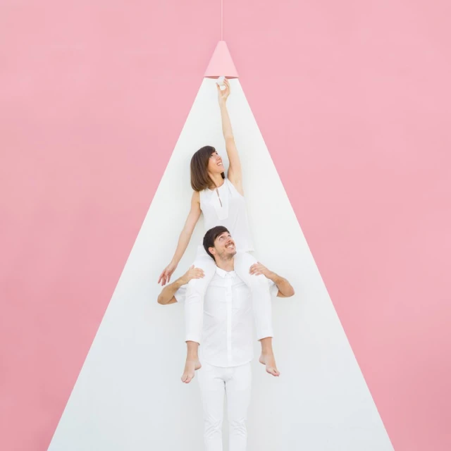 Woman on man's shoulders reaches for bulb, both wearing white, against a pink and white geometric backdrop, lit by hanging lamp.