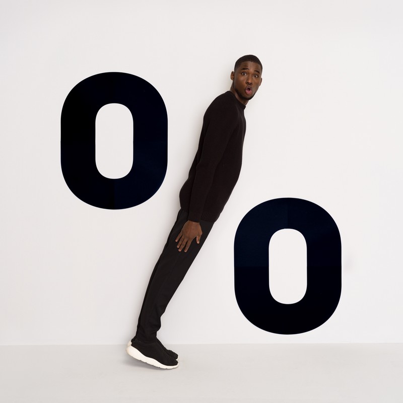 A Black man in a black sweater and pants strikes a pose in front of the two black '0' symbols on the white background. The man has a surprised expression.