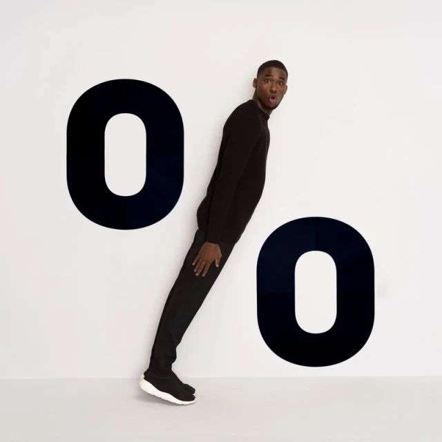 A Black man in a black sweater and pants strikes a pose in front of the two black '0' symbols on the white background. The man has a surprised expression.