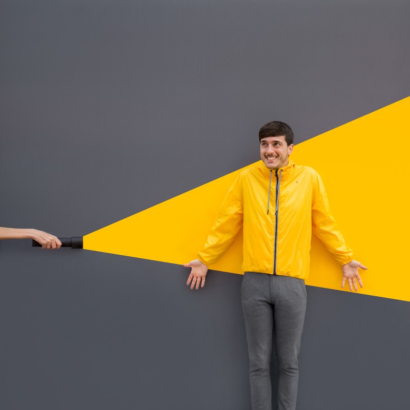 Smiling man in yellow jacket with outstretched arms, illuminated by flashlight against grey and yellow wall, creating a playful scene.