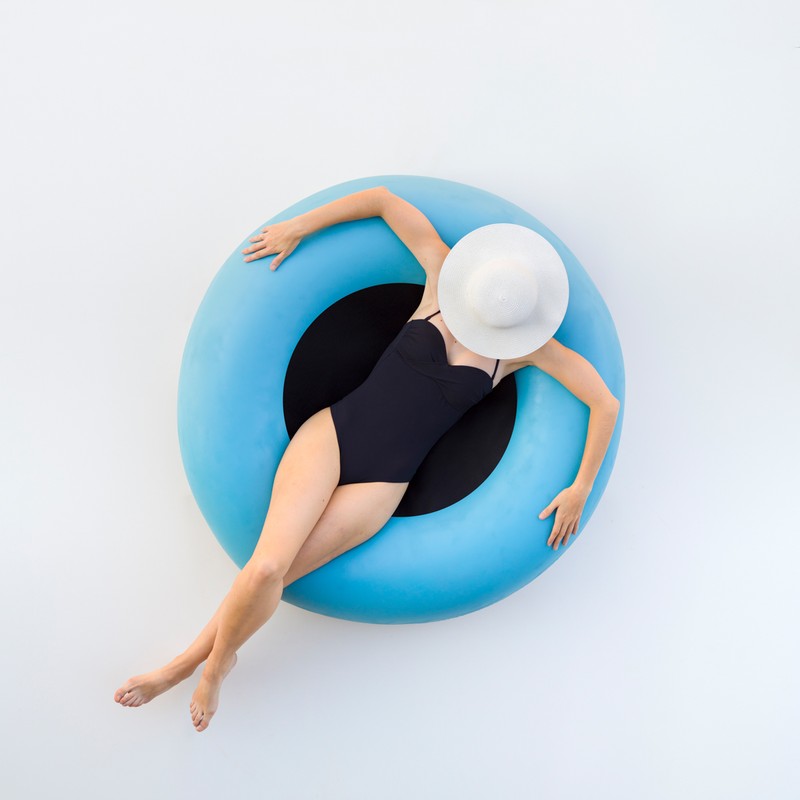 Aerial shot of a woman in a black bathing suit and white hat floating in a light blue inner tube against a white background.
