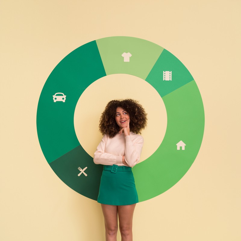 Young adult woman with curly hair, wearing a skirt and blouse, stands in front of a green circular diagram against a light yellow background.