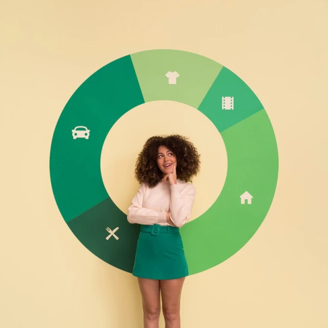 Young adult woman with curly hair, wearing a skirt and blouse, stands in front of a green circular diagram against a light yellow background.