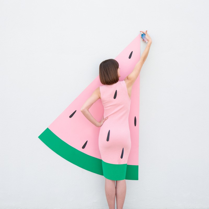 Woman in watermelon dress takes a selfie in front of a white wall. Playful fashion and creative self-portrait with bright colors.