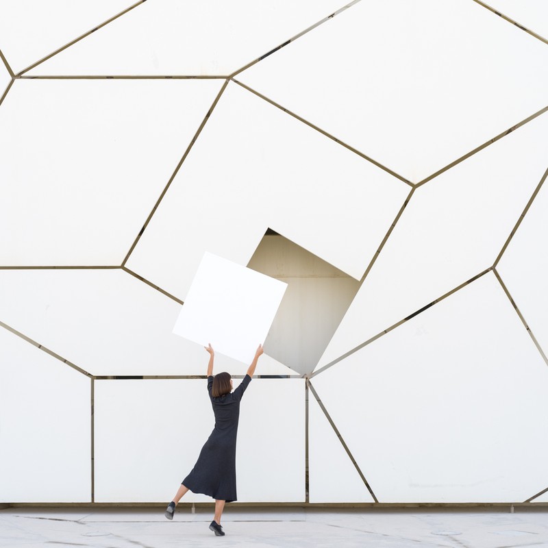 A woman in a dark dress stands in front of a white geometric wall holding a white square against the patterned background indoors.