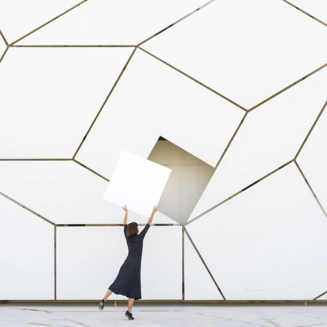 A woman in a dark dress stands in front of a white geometric wall holding a white square against the patterned background indoors.