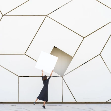 A woman in a dark dress stands in front of a white geometric wall holding a white square against the patterned background indoors.