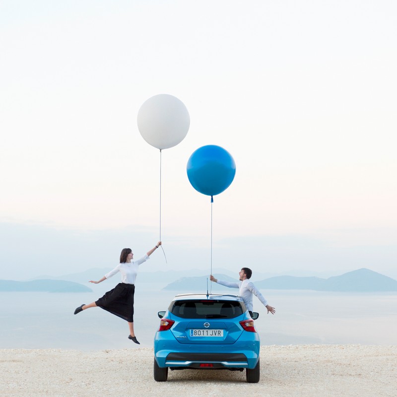 Couple playfully poses with blue car and oversized balloons against a scenic backdrop of water and mountains, creating a whimsical and lighthearted image.