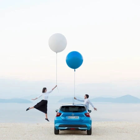 Couple playfully poses with blue car and oversized balloons against a scenic backdrop of water and mountains, creating a whimsical and lighthearted image.