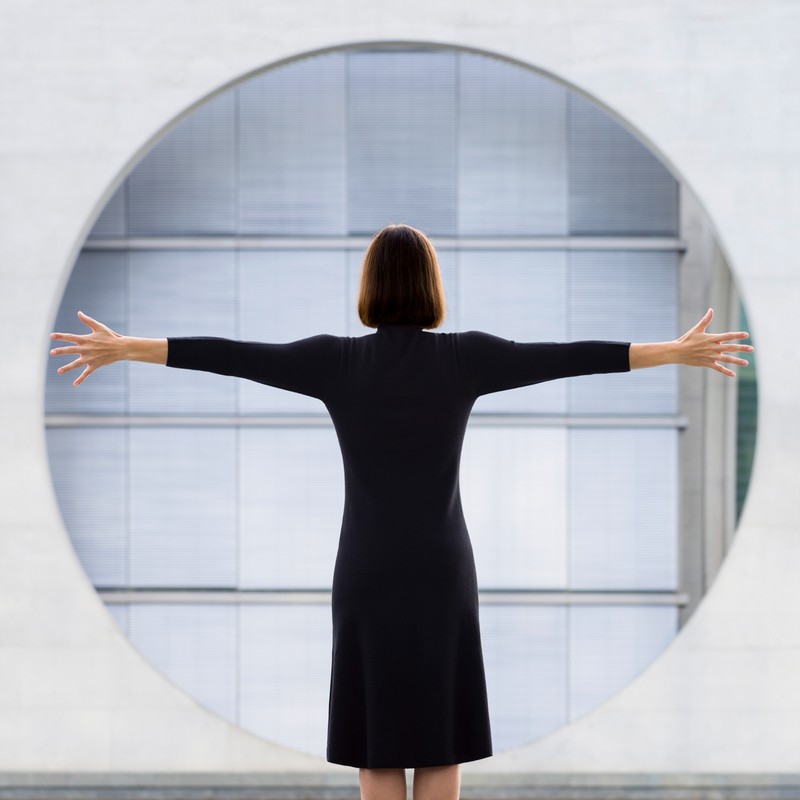 Back view of woman in black dress with arms outstretched in front of round window, architectural setting with neutral colors.