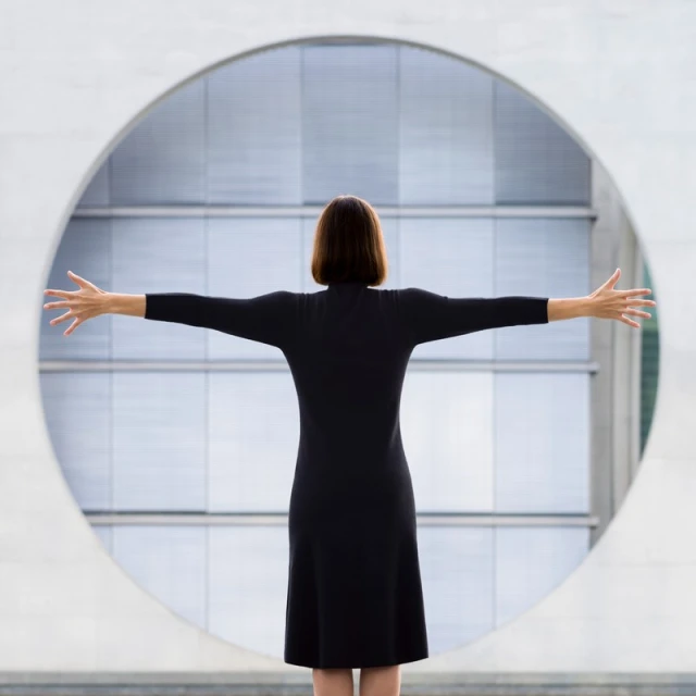 Back view of woman in black dress with arms outstretched in front of round window, architectural setting with neutral colors.
