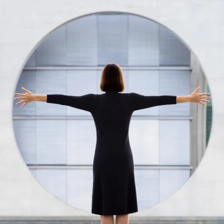 Back view of woman in black dress with arms outstretched in front of round window, architectural setting with neutral colors.
