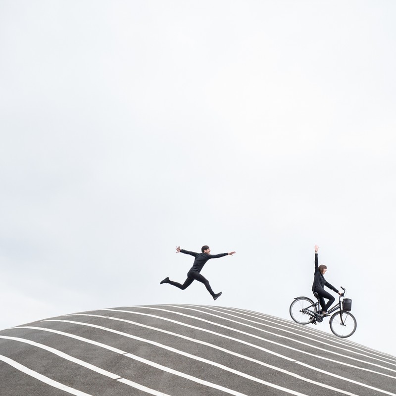 Two people in black suits pose on a striped, curved structure against a bright sky: one jumping, one on a bike with raised arm.