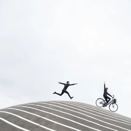 Two people in black suits pose on a striped, curved structure against a bright sky: one jumping, one on a bike with raised arm.