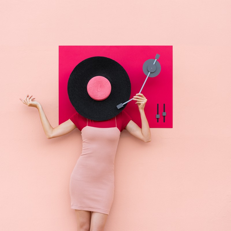 Conceptual fashion shot: female torso in pink dress, arms raised, record player as head against a peach background, pop-art vibe.