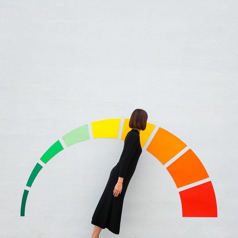 A woman in a black dress stands by a colorful, curved meter graphic, shot in a minimalist studio setting with a flat lay perspective.