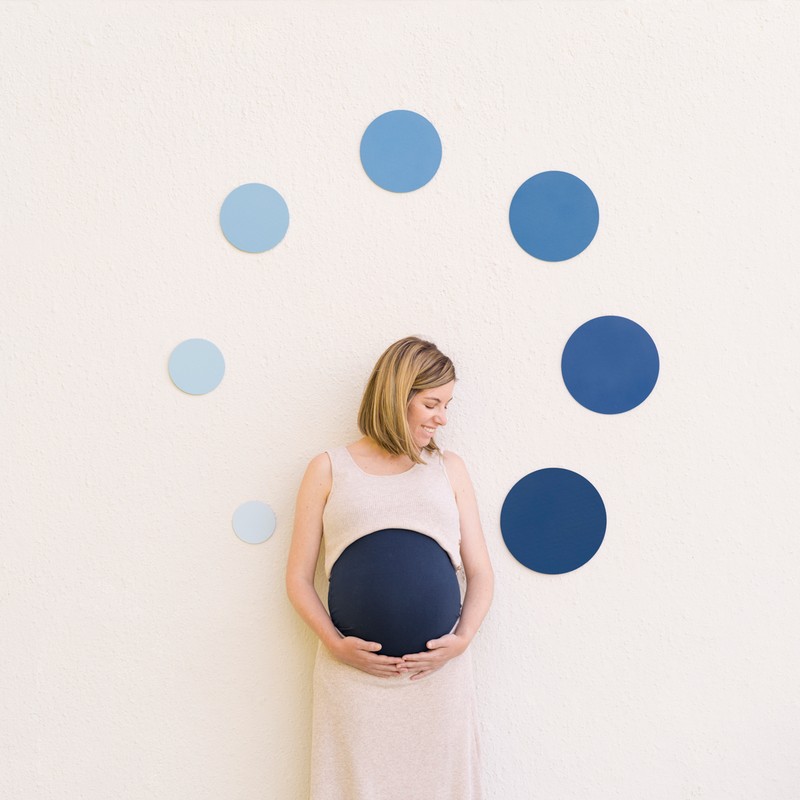 Pregnant woman in a beige dress holds her belly, which is covered in a navy fabric circle, posing in front of a wall with blue circles.