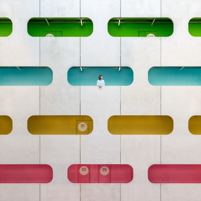 Architectural shot of a woman standing in a blue balcony nook, viewed from the front, in a grid of colorful rectangular openings.