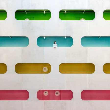 Architectural shot of a woman standing in a blue balcony nook, viewed from the front, in a grid of colorful rectangular openings.