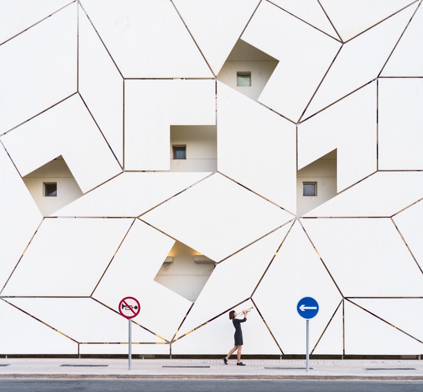 Woman in black dress playing trumpet on sidewalk against geometric patterned wall with windows and street signs. Architecture.