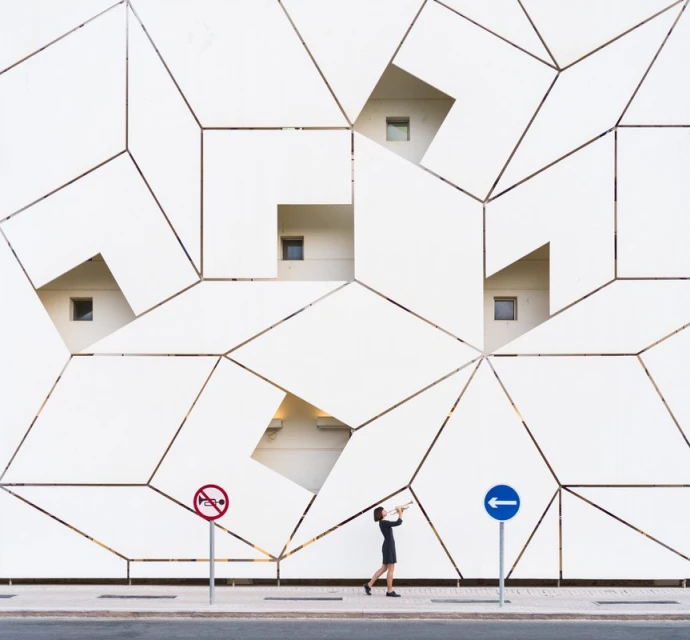 Woman in black dress playing trumpet on sidewalk against geometric patterned wall with windows and street signs. Architecture.