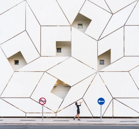 Woman in black dress playing trumpet on sidewalk against geometric patterned wall with windows and street signs. Architecture.