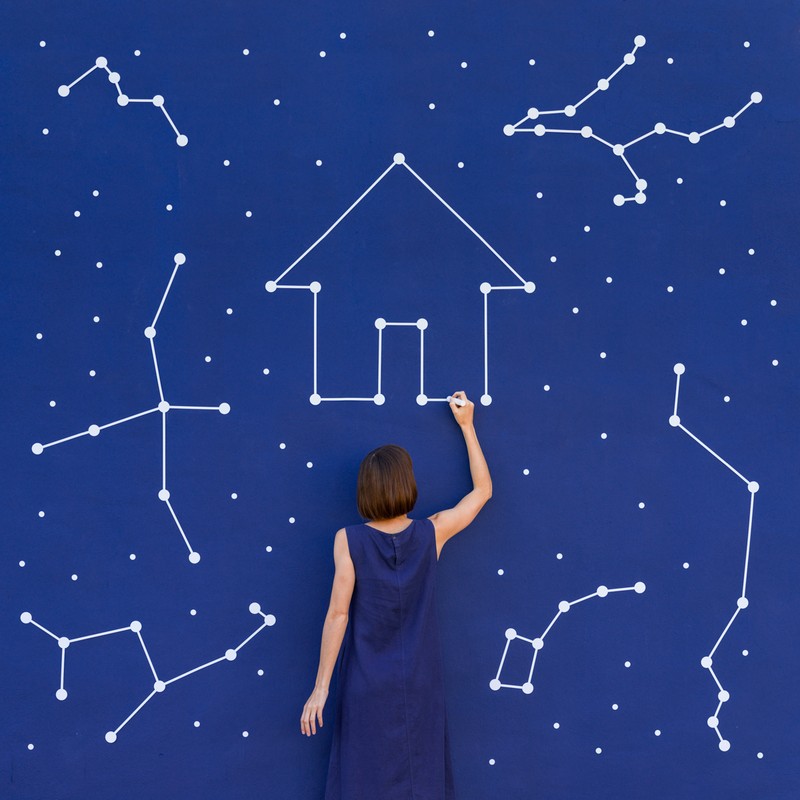 Back view of a woman drawing a house with constellations on a blue wall, creating a whimsical, celestial real estate concept.